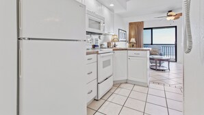 A modern kitchen with white cabinets, a microwave, and a refrigerator. There is a dining area with a table and chairs, and a view of the outside through a large window. - Beacher's Oceanfront Balcony Suite with Onsite Front desk and staff (St. Augustine)