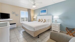 A hotel room with a bed, a TV, a ceiling fan, a window with curtains, a nightstand, and a chair. - Beacher's Oceanfront Balcony Suite with Onsite Front desk and staff (St. Augustine)