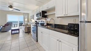 A modern kitchen with white cabinets, a black countertop, and a microwave. There is a dining area with a table and chairs, and a balcony with a view. - Beacher's Oceanfront Balcony Suite with Onsite Front desk and staff (St. Augustine)