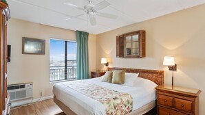 A hotel room with a large bed, two bedside lamps, a wooden nightstand, a ceiling fan, a window with curtains, and a wall-mounted air conditioning unit.