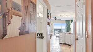 A narrow hallway leading to a kitchen with white cabinets, a tiled floor, and a view of the ocean through a window.