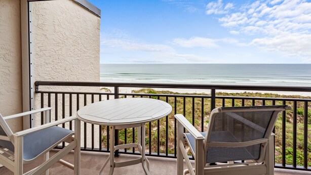 A balcony with a white table and two chairs, overlooking a beach with the ocean and a clear sky.