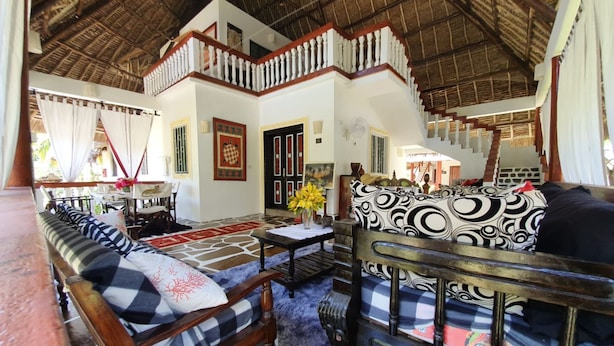 A spacious living area with a high ceiling and a staircase leading to an upper level. There is a sofa and a chair with patterned cushions, a small table with a vase of flowers, and a dining area with a table and chairs. - Nuru Ya Erika Villas (Malindi)