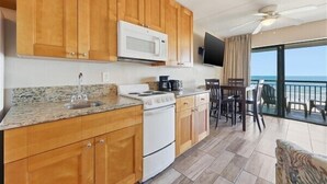 A kitchen with wooden cabinets, a microwave, a stove, and a sink. There is a dining area with chairs and a table. The room has a view of the ocean. - Your New Favorite Beachside Escape | Top Floor & Oceanfront (St. Augustine)
