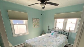 Un dormitorio con cama, ventilador de techo, dos ventanas y una pared con un letrero decorativo.