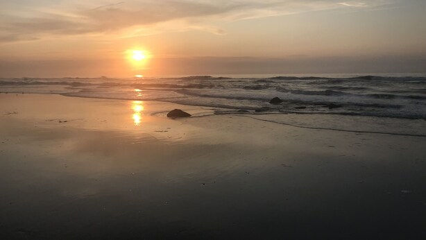 Una playa con el sol poniéndose sobre el océano, olas rompiendo en la orilla y un cielo despejado.