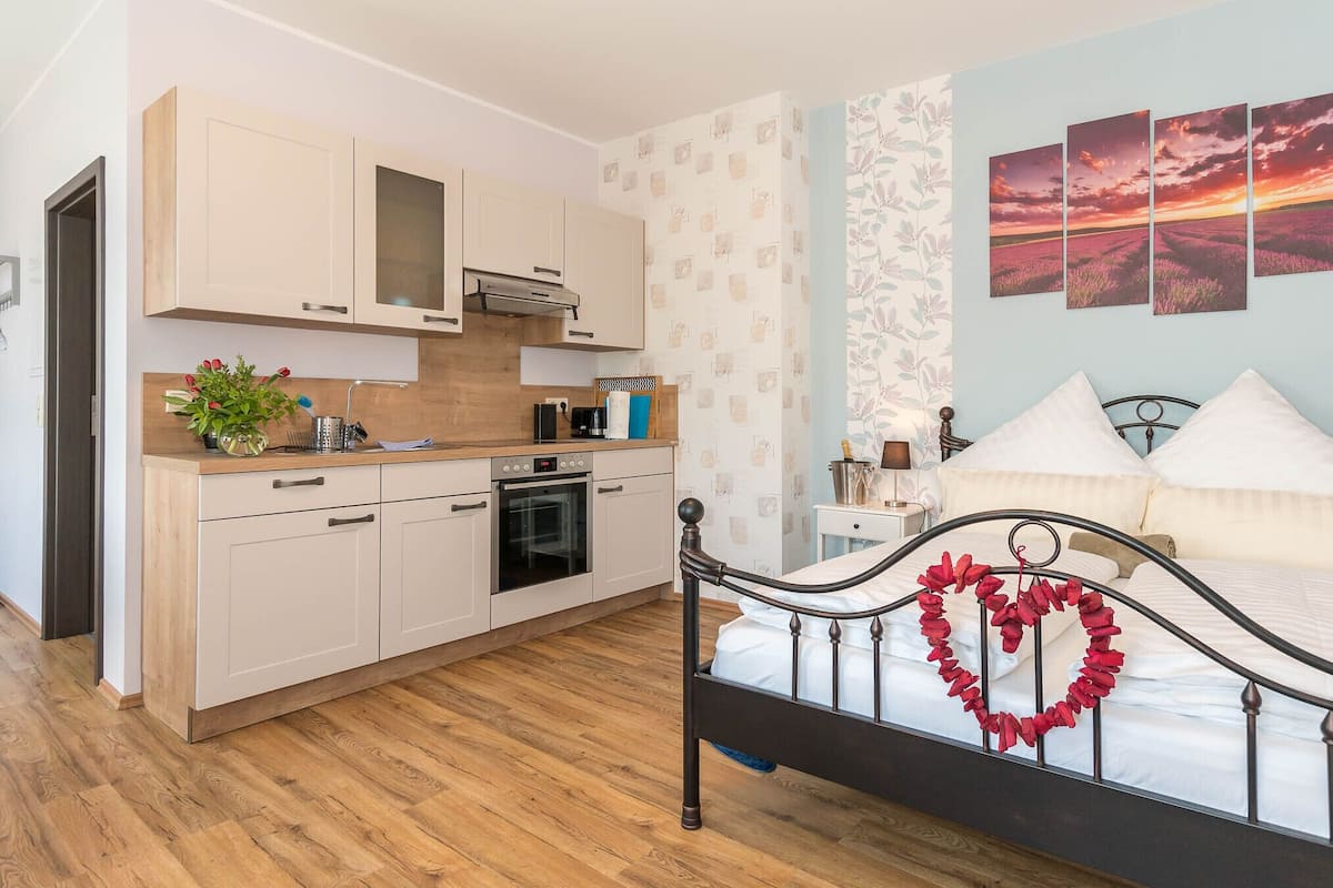 Un dormitorio con cama, mesita de noche, alacena y una cocina con armarios y estufa. En la pared, un cuadro con un atardecer.