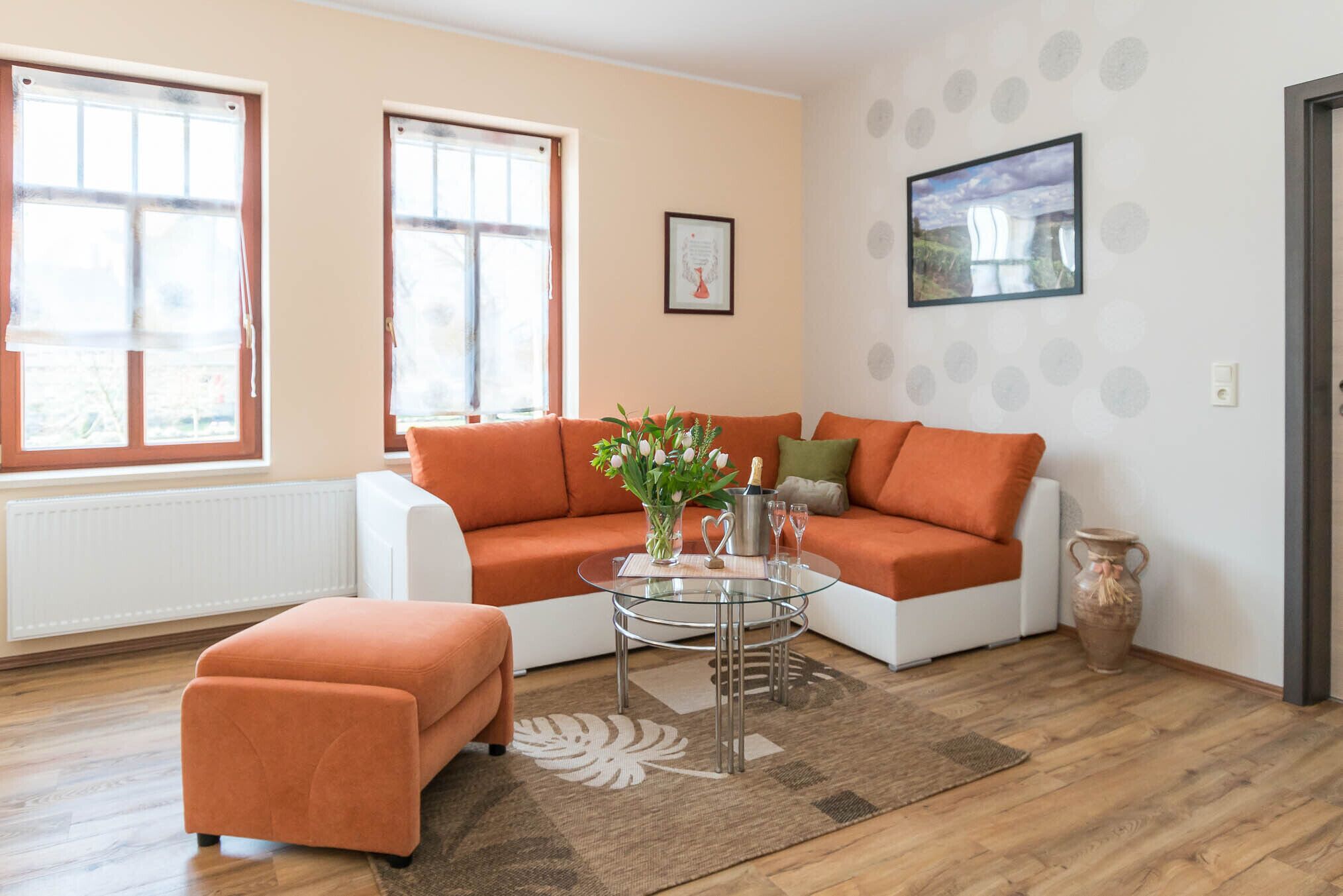 Una sala de estar con un sofá blanco, cojines naranjas, una mesa de centro de vidrio y un jarrón con flores.