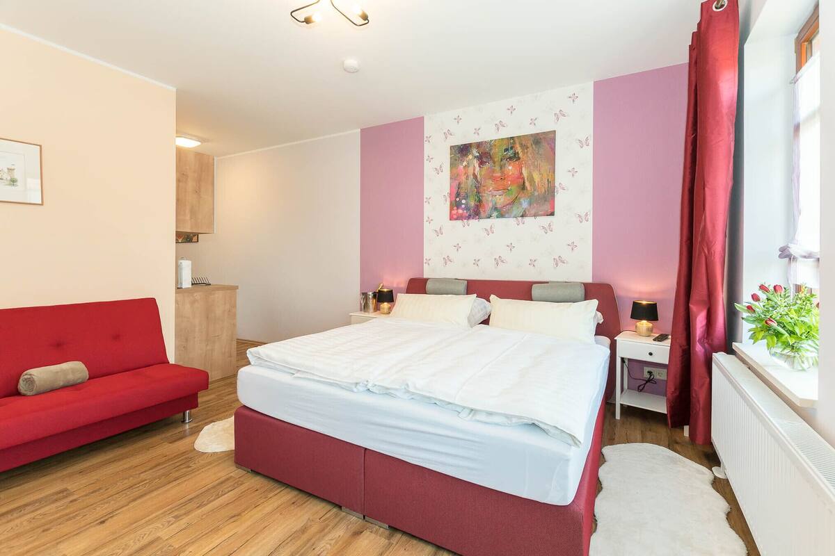 Un dormitorio con cama, sofá rojo, mesita de noche y ventana con cortinas.