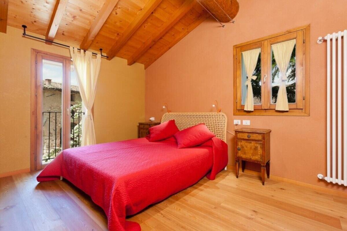 A bedroom with a wooden ceiling, a bed with red bedding, a small wooden nightstand, and a window with curtains.