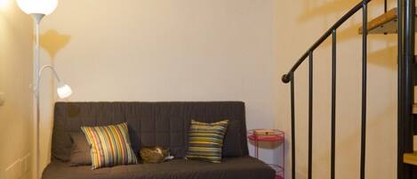 A dark-colored sofa with striped and solid pillows, a floor lamp, and a small pink side table with a red object.
