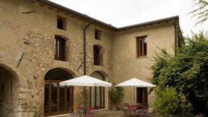 A stone building with arched doorways and windows, outdoor seating with umbrellas, and a garden area with rocks and greenery. - Solimago Dimora Storica (Solferino)