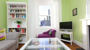 A living room with a bookshelf, a purple armchair, a glass coffee table, a television, and a fireplace.
