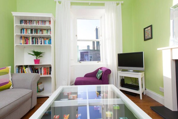 A living room with a bookshelf, a purple armchair, a glass coffee table, a television, and a fireplace.