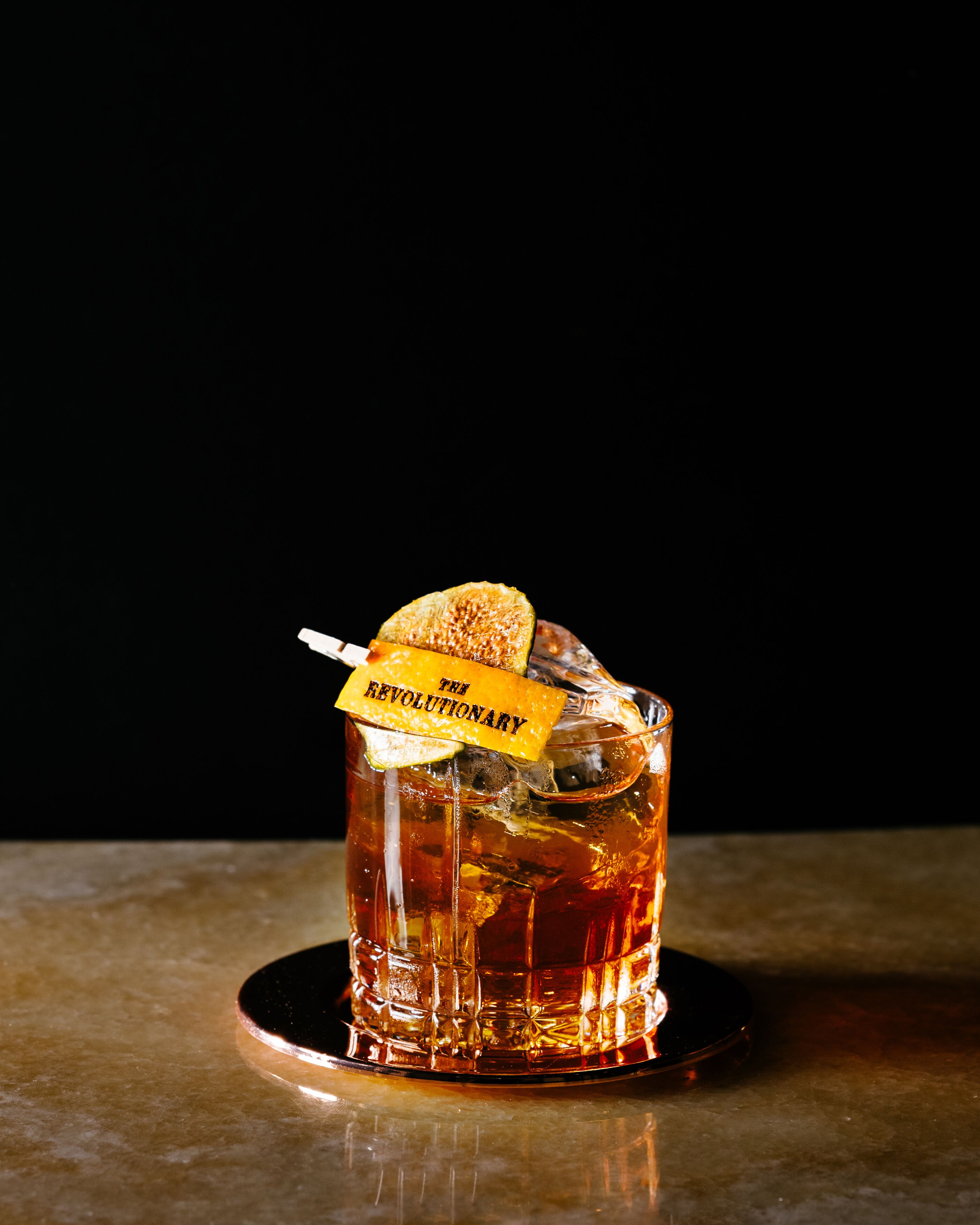 A glass of amber-colored beverage with a garnish and a label that reads "THE REVOLUTIONARY" on a coaster.