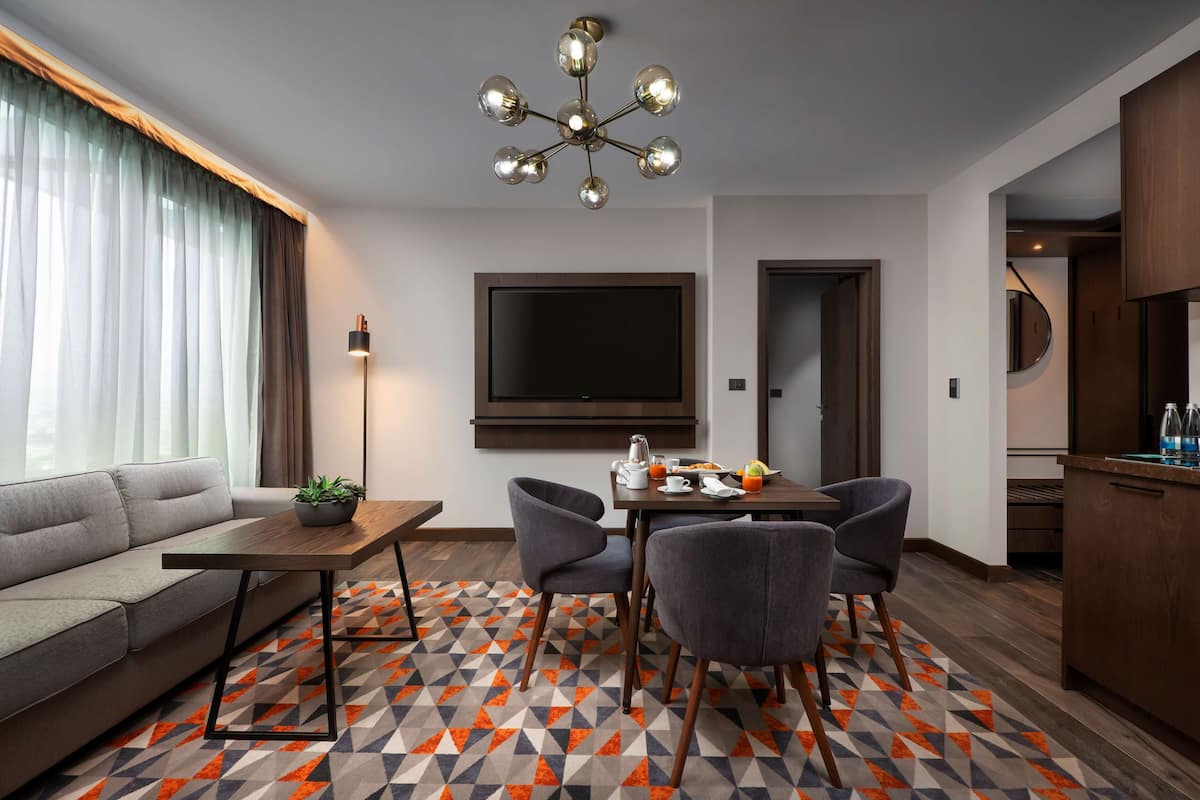 Ein modernes Hotelzimmer mit einer Couch, einem Esstisch, Stühlen, einem Fernseher und einem gemusterten Teppich.