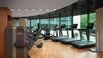 A modern gym with cardio equipment, a wooden floor, and large windows offering a view of greenery.