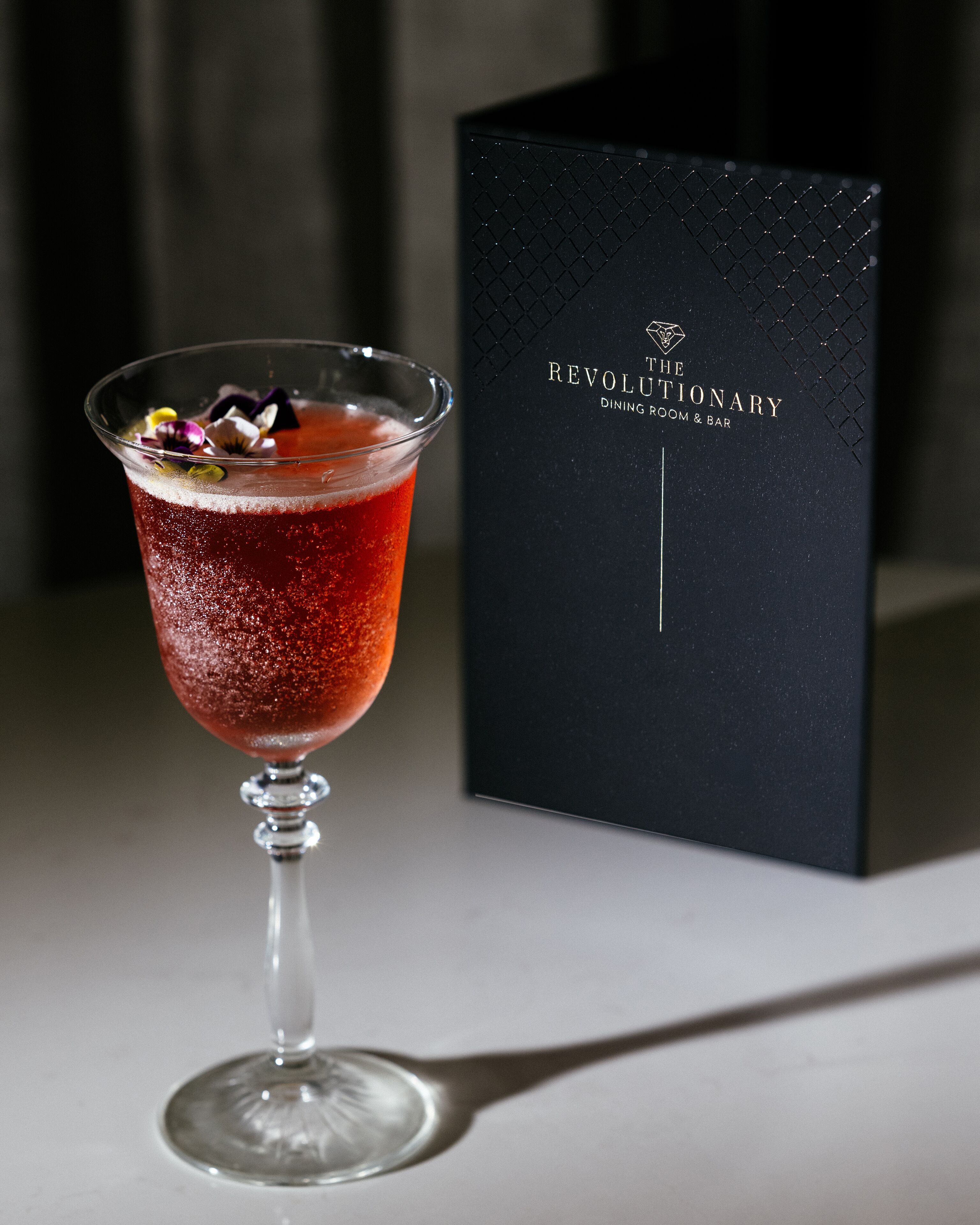 A cocktail in a martini glass with a decorative garnish, placed on a table next to a black box with the text "The Revolutionary Dining Room & Bar".