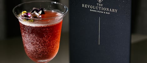 A cocktail in a martini glass with a decorative garnish, placed on a table next to a black box with the text "The Revolutionary Dining Room & Bar".