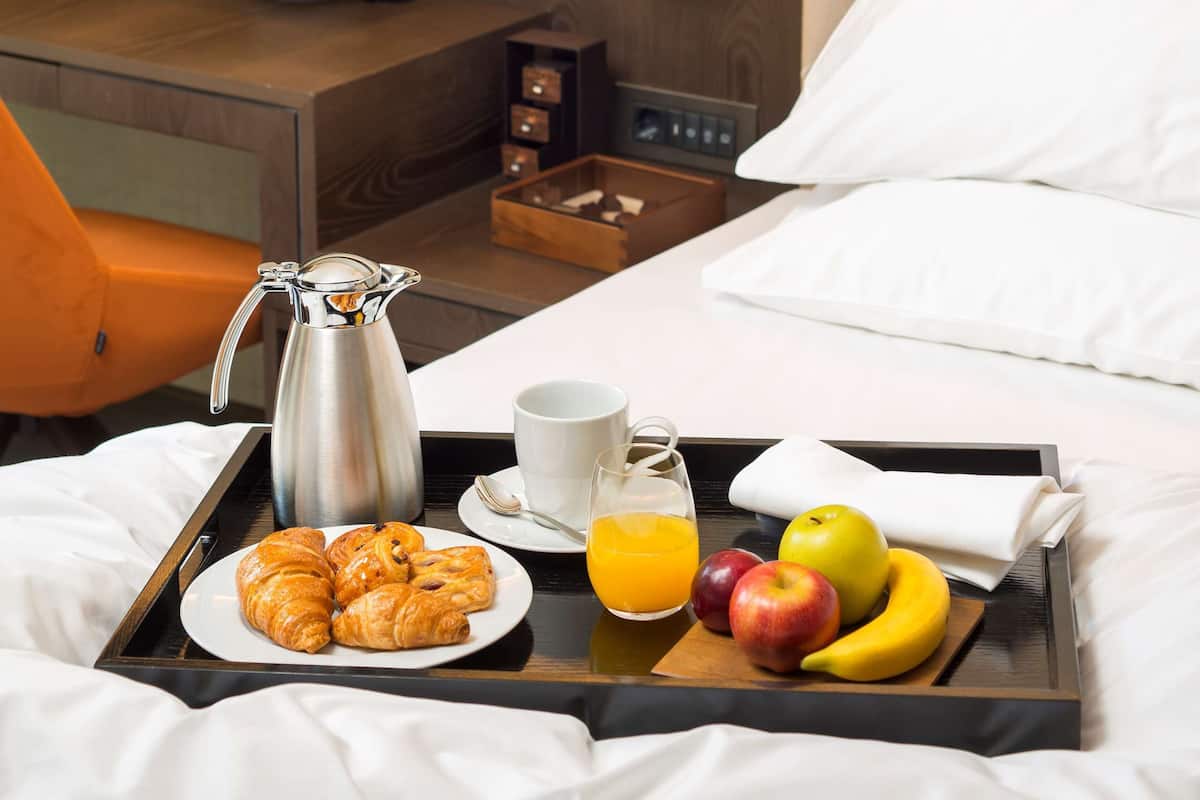 Ein ordentlich bezogenes Bett mit einem Tablett mit Frühstücksartikeln, darunter Croissants, Früchte und Saft, sowie ein Nachttisch mit Pflanze und Büchern.