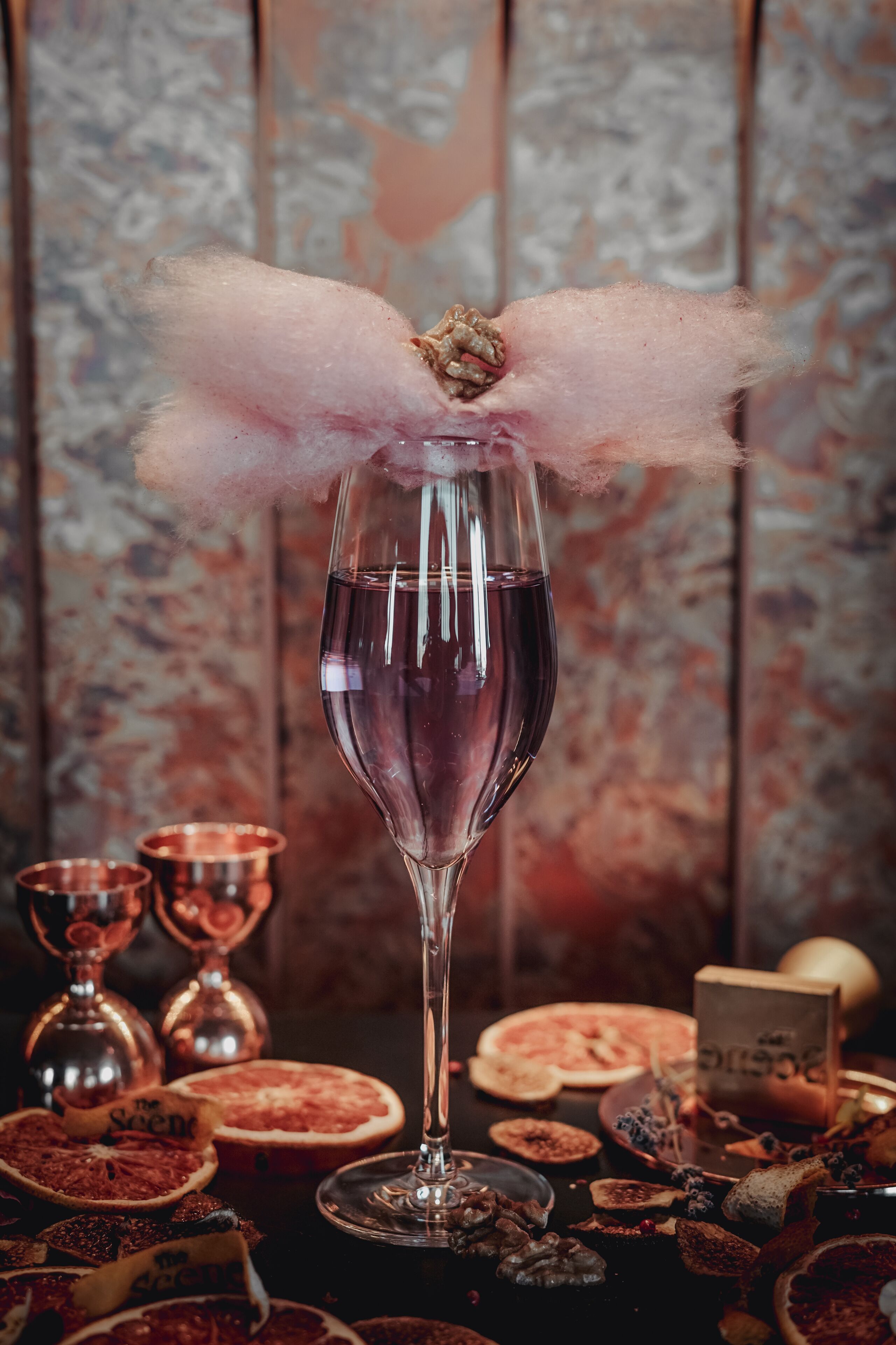 Ein Glas roter Wein mit einer rosa, flauschigen Dekoration, umgeben von verschiedenen Speisen und Kupfergefäßen.