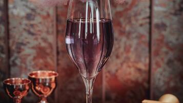 Una copa de vino tinto con una decoración rosada y esponjosa en la parte superior, rodeada de diversos alimentos y recipientes de cobre.