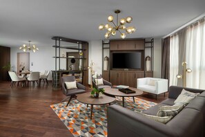 A modern living room with a sofa, armchairs, a coffee table, and a TV. There is a dining area with a table and chairs, and a kitchen area with cabinets and a TV.