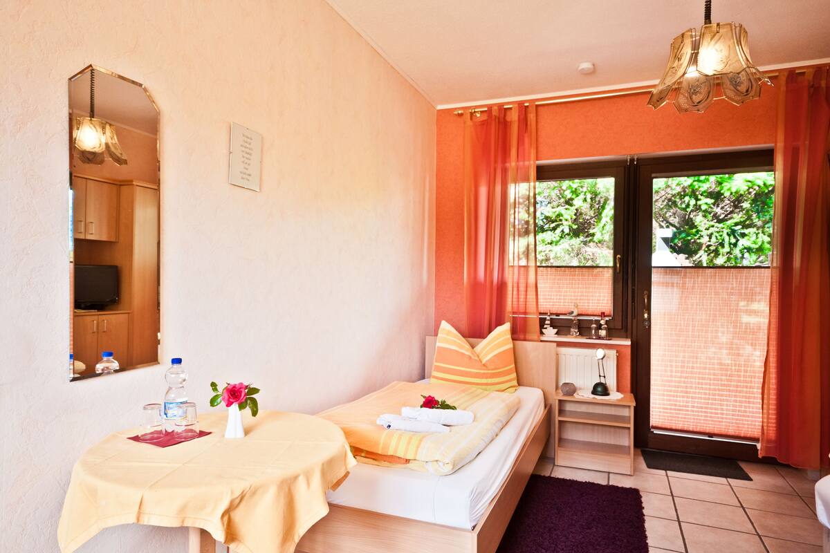 A hotel room with a bed, a small table with a tablecloth, a mirror, a nightstand, a lamp, and a view of greenery outside.