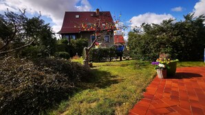 A house with a red-tiled roof, surrounded by a garden with a paved walkway. - Pension Fischerhaus (Trassenheide)