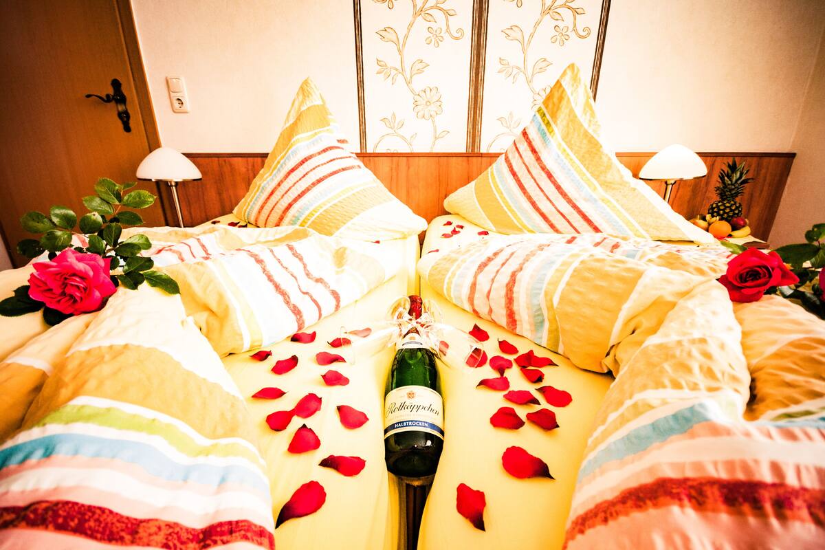 A bed with a bottle of champagne, rose petals, and a red rose.