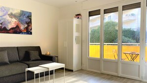 A modern living room with a grey sofa, a white coffee table, and a large window with a view of a yellow fence and greenery. - Large and Quiet Studio with Balcony (Cannes)