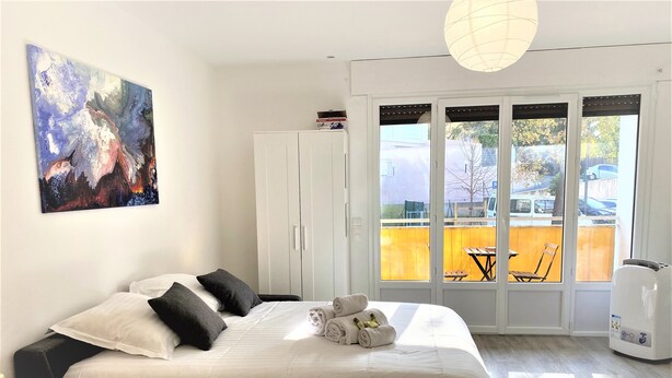 A modern hotel room with a large bed, a white wardrobe, a painting on the wall, and a view of the outside through a glass door. - Large and Quiet Studio with Balcony (Cannes)
