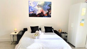 A neatly made bed with white linens, two black pillows, and a rolled towel. There is a painting above the bed, a nightstand with a lamp, and a wardrobe to the right. - Large and Quiet Studio with Balcony (Cannes)