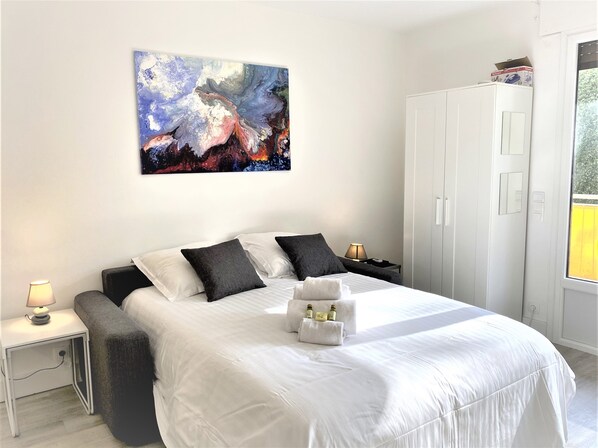 A modern bedroom with a large bed, white walls, a painting, a wardrobe, and a bedside table with a lamp. - Large and Quiet Studio with Balcony (Cannes)