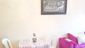 Uma pequena área de refeições com uma mesa coberta por uma toalha estampada, duas cadeiras brancas, um frasco decorativo e uma obra de arte com caligrafia árabe emoldurada na parede.