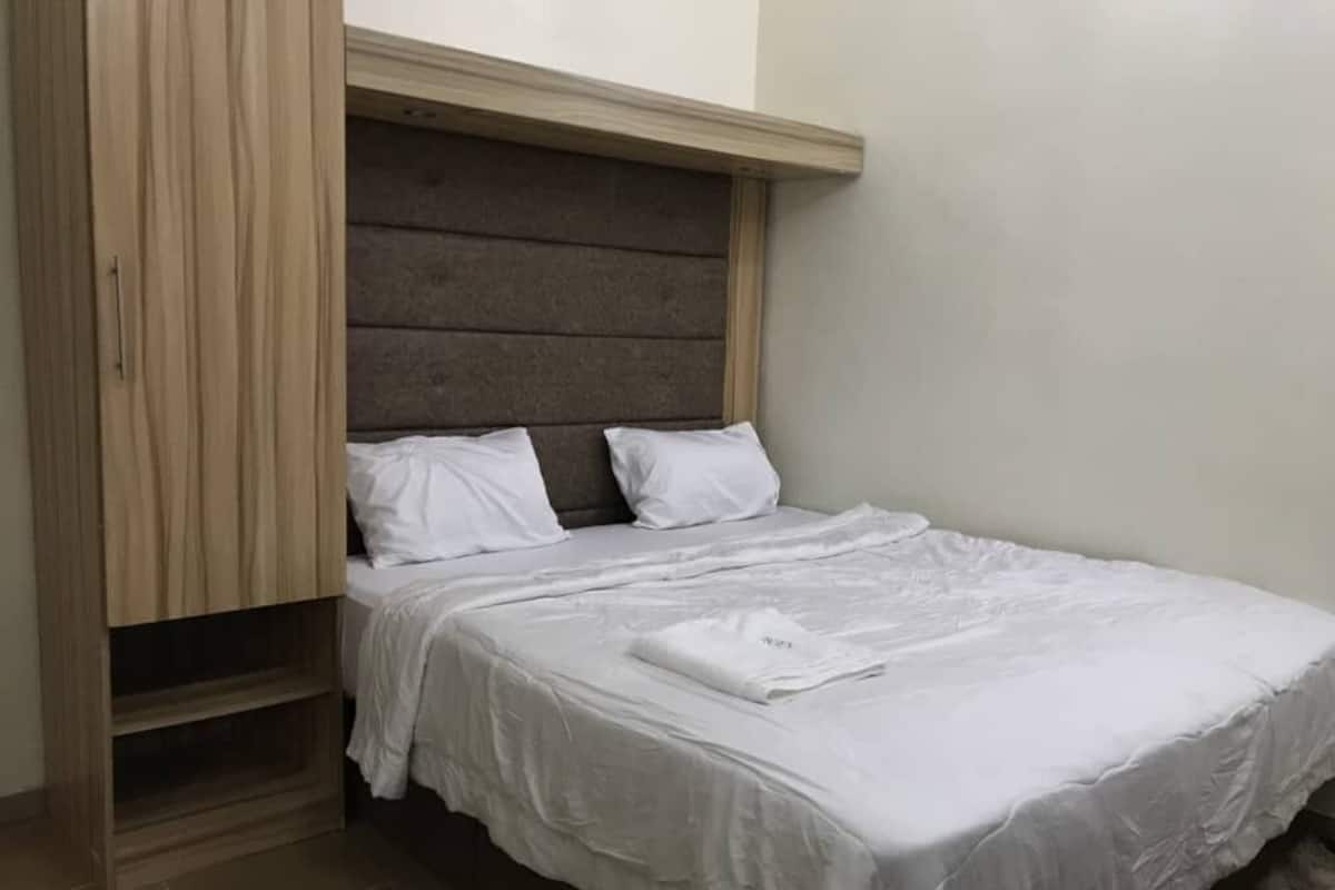 A single bed with a wooden headboard, white bedding, and a small shelf unit to the left.