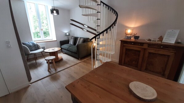 A modern living room with a wooden table, a spiral staircase, a sofa, and a window with a view of greenery.