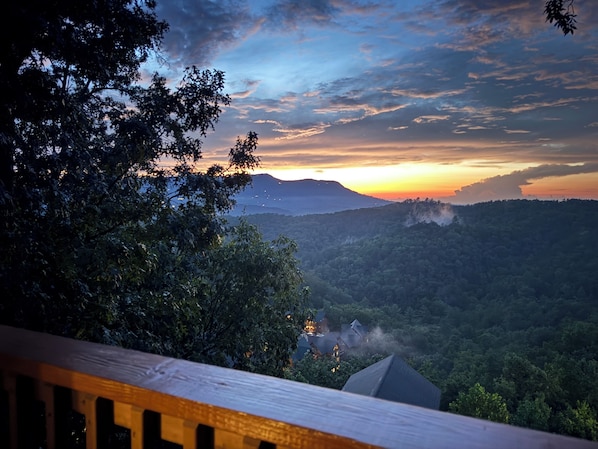 A view from a balcony overlooking a forested landscape with a sunset in the background.