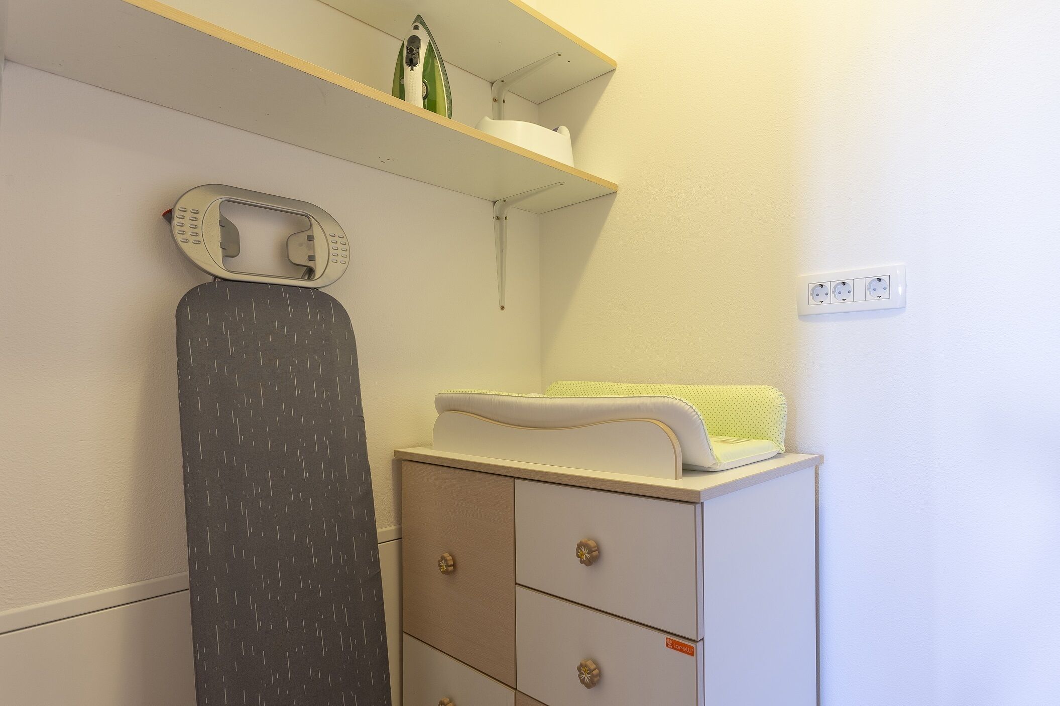 A neatly organized room with a white dresser, a shelf with a kettle and a container, and a power outlet on the wall.