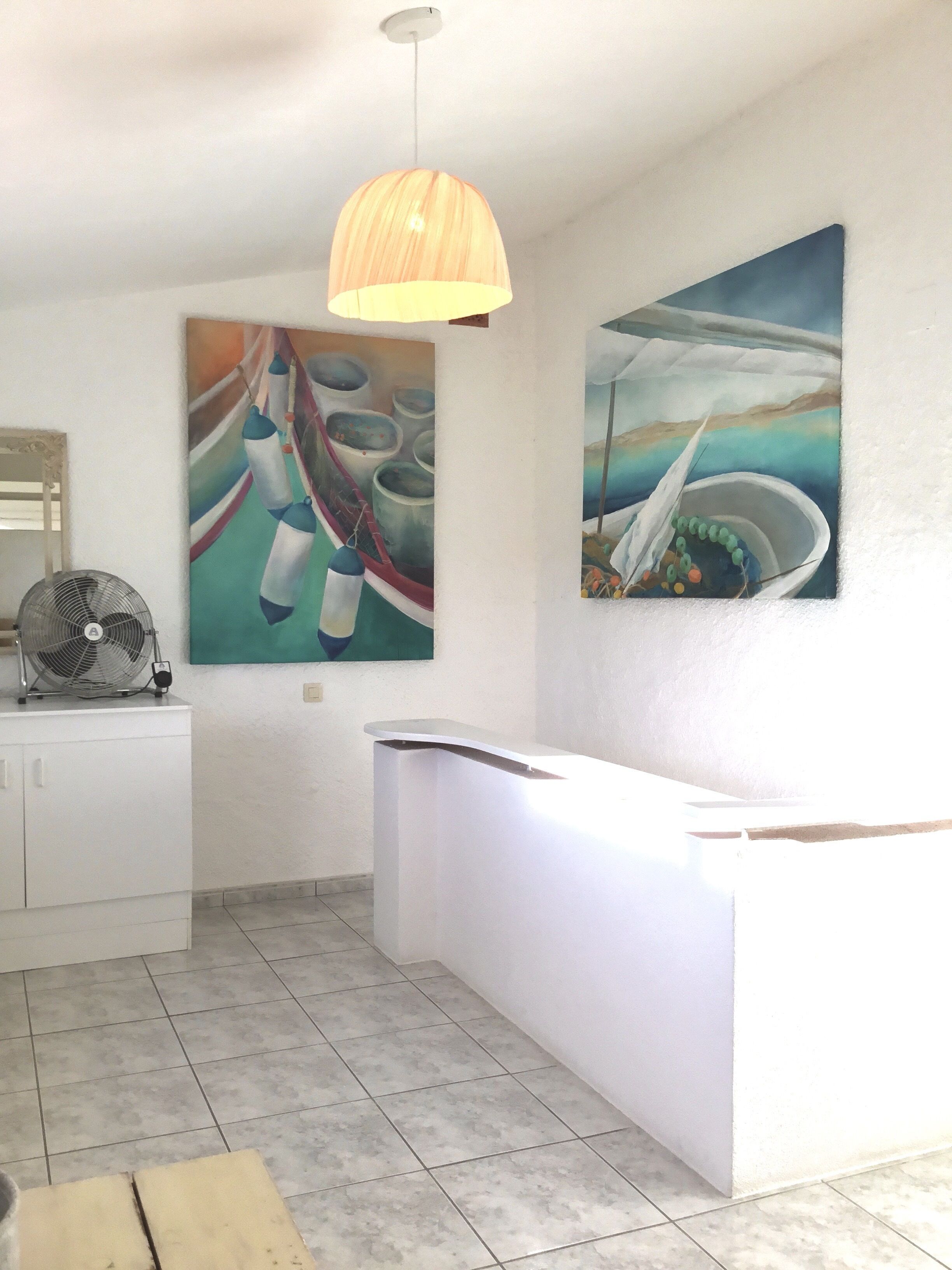 A modern reception area with a white counter, a ceiling fan, and two paintings on the wall.