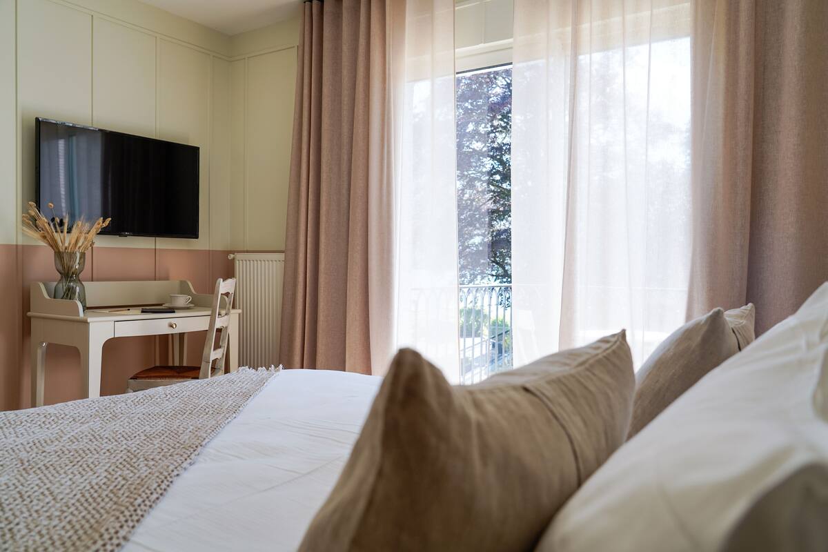 A bedroom with a bed, a desk, a chair, a TV, and a window with curtains.