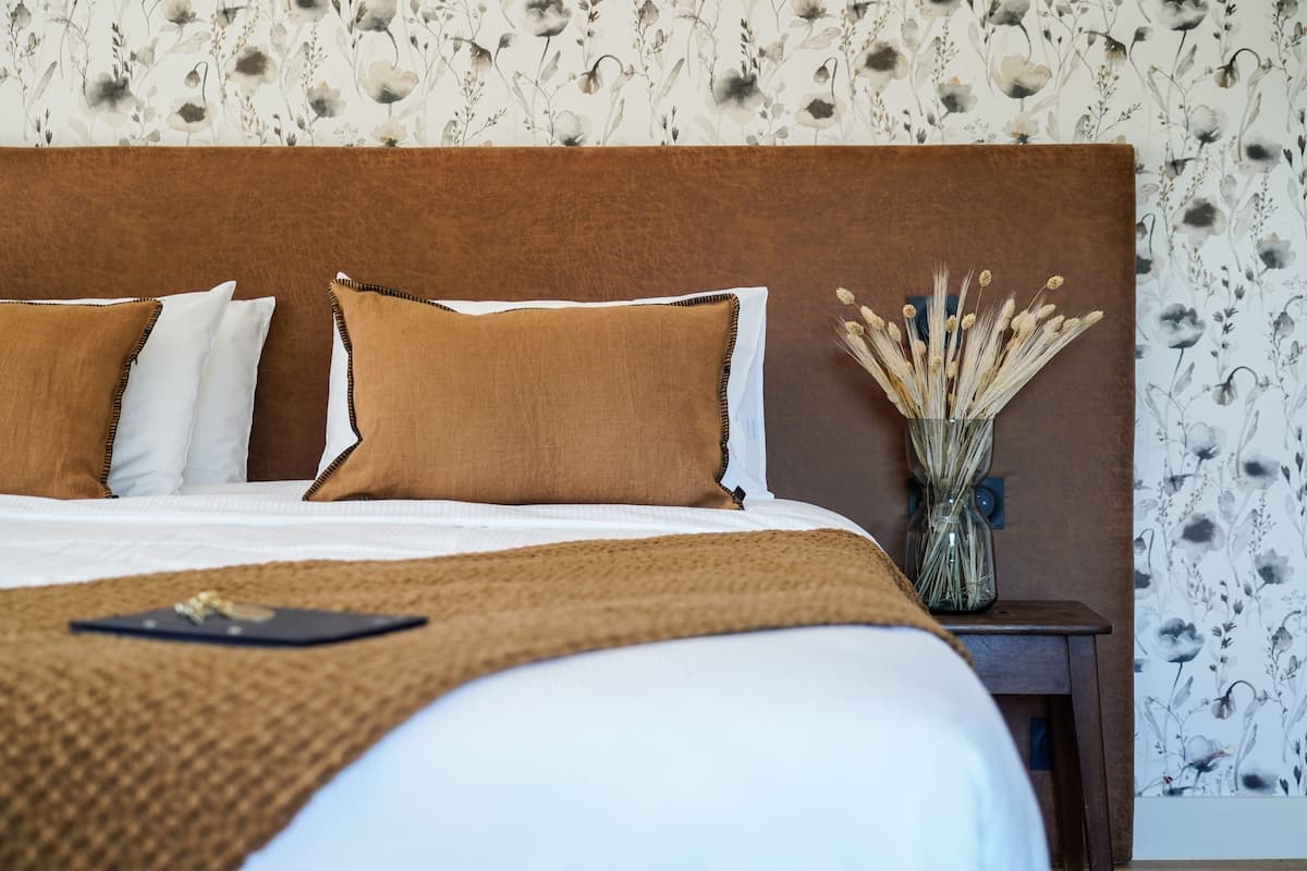 A neatly made bed with a brown headboard, white and brown pillows, and a brown throw blanket. A small table with a vase of dried flowers is beside the bed.