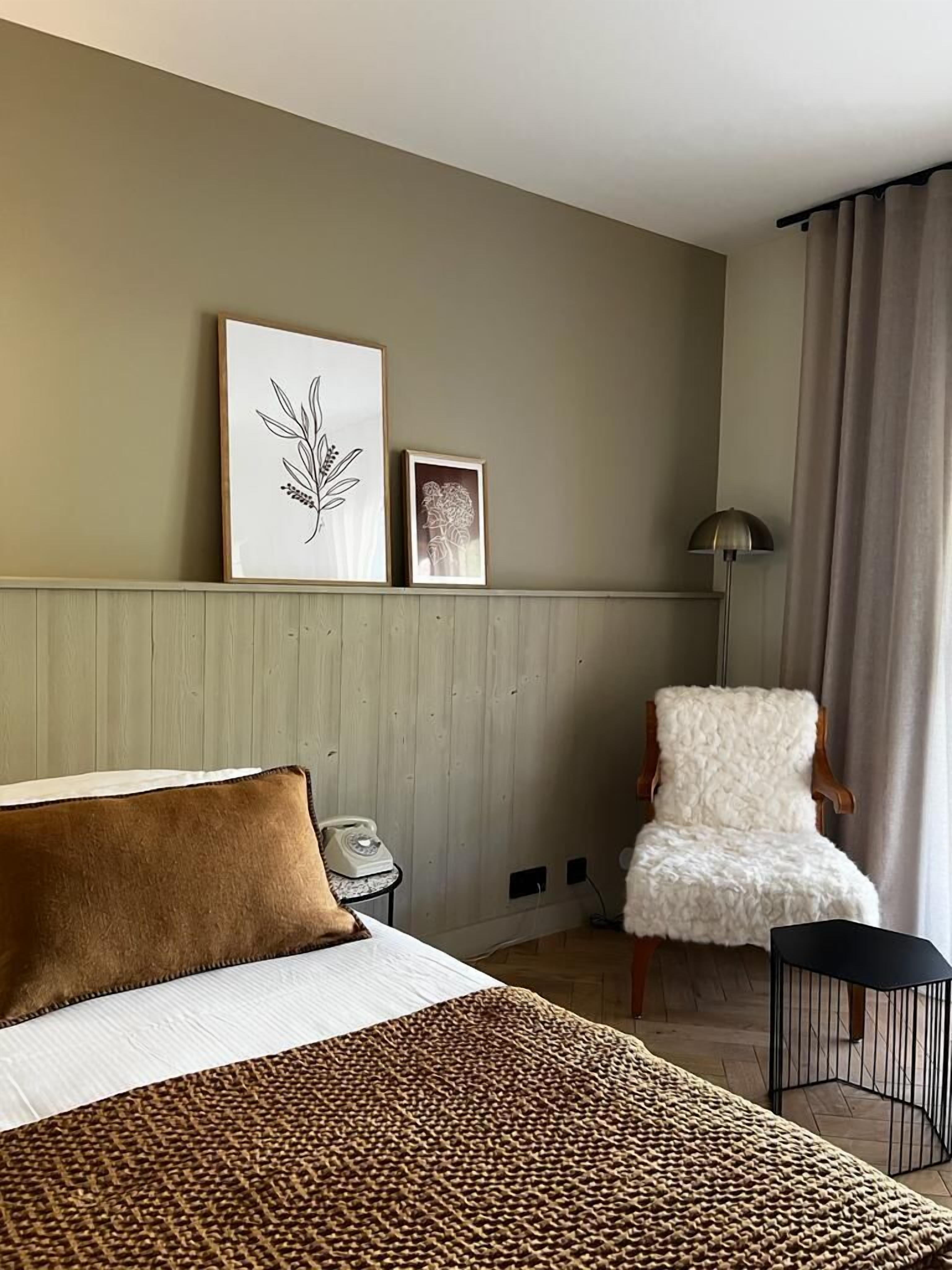 A hotel room with a bed, a chair, a lamp, and two framed pictures on the wall.