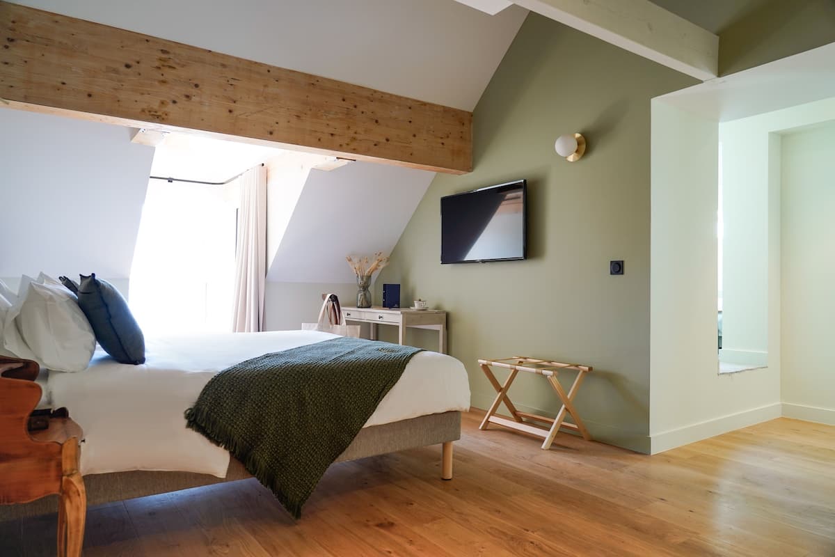 A bedroom with a bed, wooden floor, a desk, a chair, and a TV on the wall.