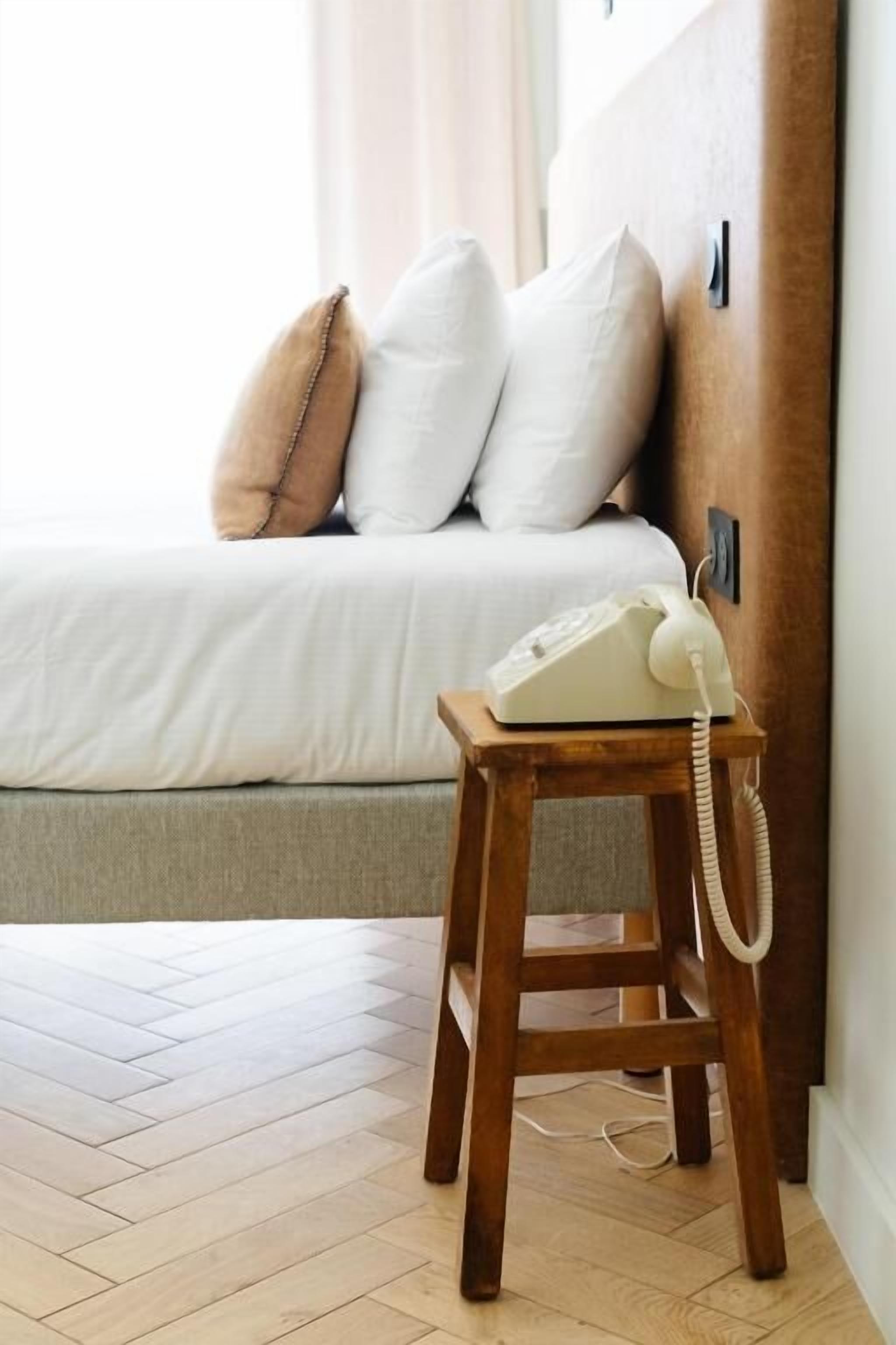 Une table de chevet en bois avec un téléphone blanc, un lit avec une literie blanche et deux oreillers, et un plancher en bois.