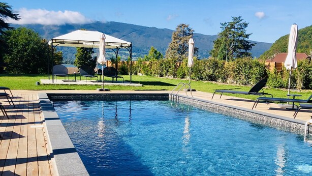 Une piscine rectangulaire avec une terrasse en bois d’un côté et un espace salon couvert avec des parasols à l’arrière-plan.