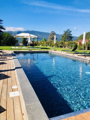 Una piscina rectangular con un deck de madera en un lado y una zona de estar techada con sombrillas al fondo.