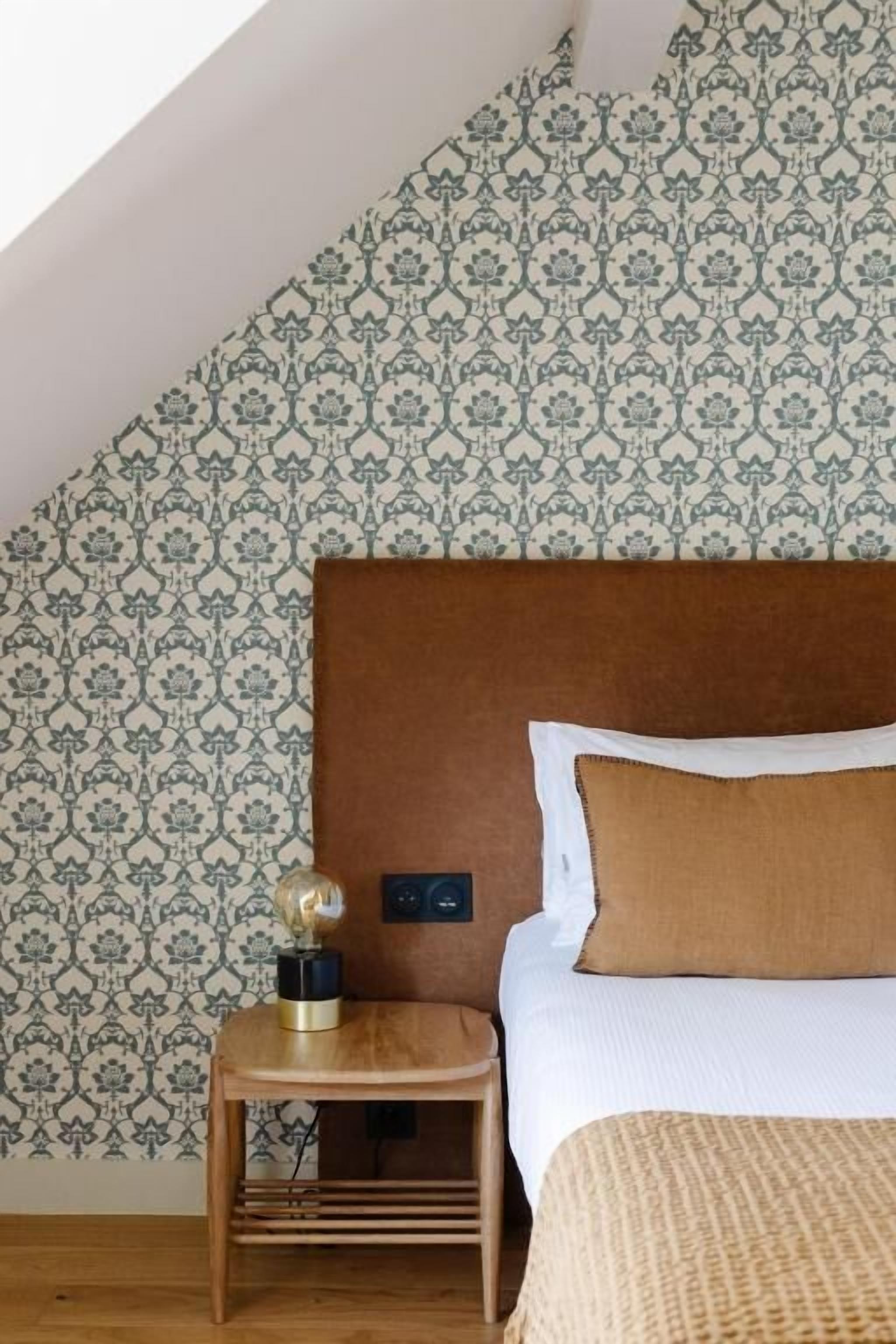 A bedroom with a patterned wallpaper, a wooden bedside table with a lamp, and a bed with a brown pillow.