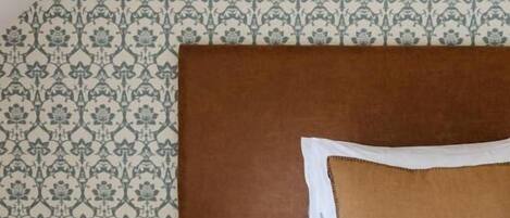 A bedroom with a patterned wallpaper, a wooden bedside table with a lamp, and a bed with a brown pillow.