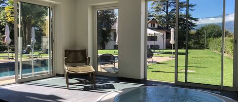 Un área de piscina interior moderna con un jacuzzi, una silla de madera y grandes puertas corredizas que dan a un patio trasero con césped y mobiliario de exterior.
